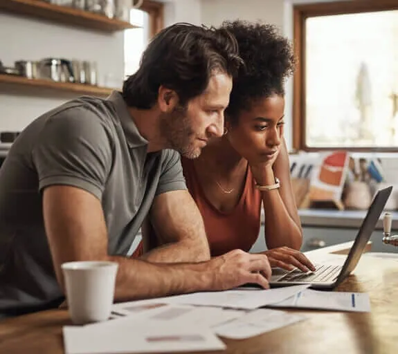 Woman with peach top is sat at her desk, smiling looking at a laptop whilst a man in a blue shirt stood next to her bends to look at the laptop, with his arm around her