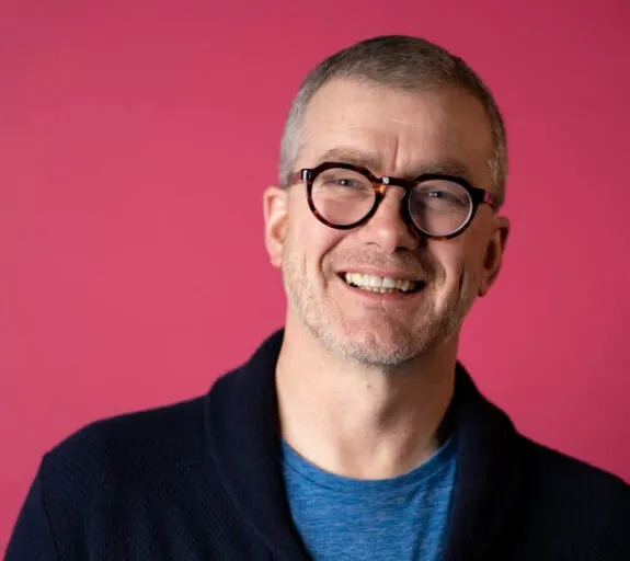 Atom bank CEO, Mark Mullen, wearing glasses and a black cardigan stood in front of a pink background, smiling