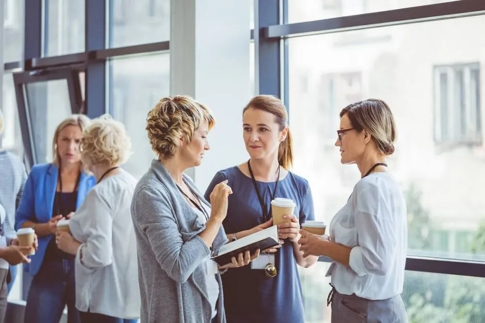 A woman holding a notepad is talking to two other women drinking coffee.