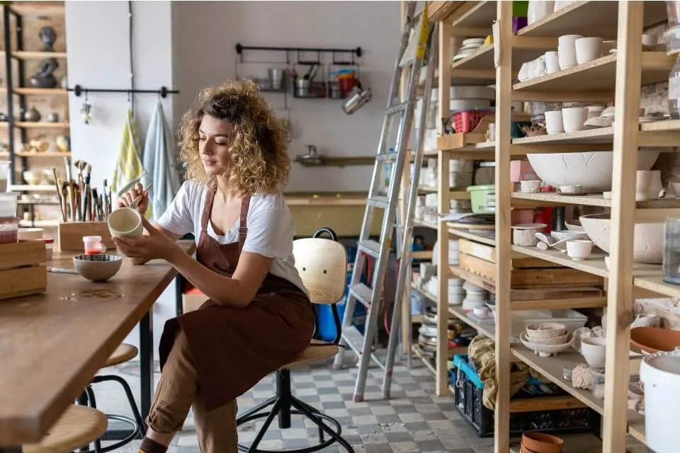 Person paints a piece of pottery, sitting in a workshop wearing an apron