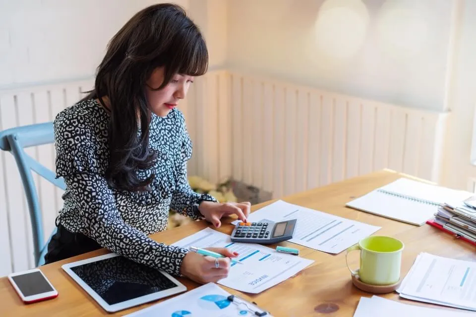 A person types into a calculator while scanning through a document, pen in hand