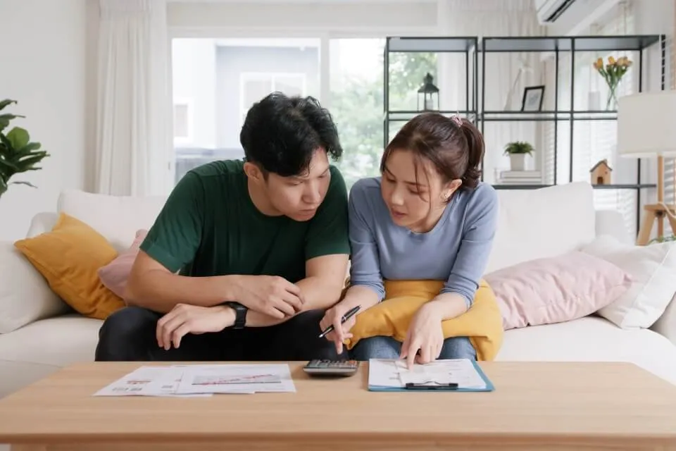 Two people, sitting together on a sofa, scan through a document on a clipboard while typing into a calculator