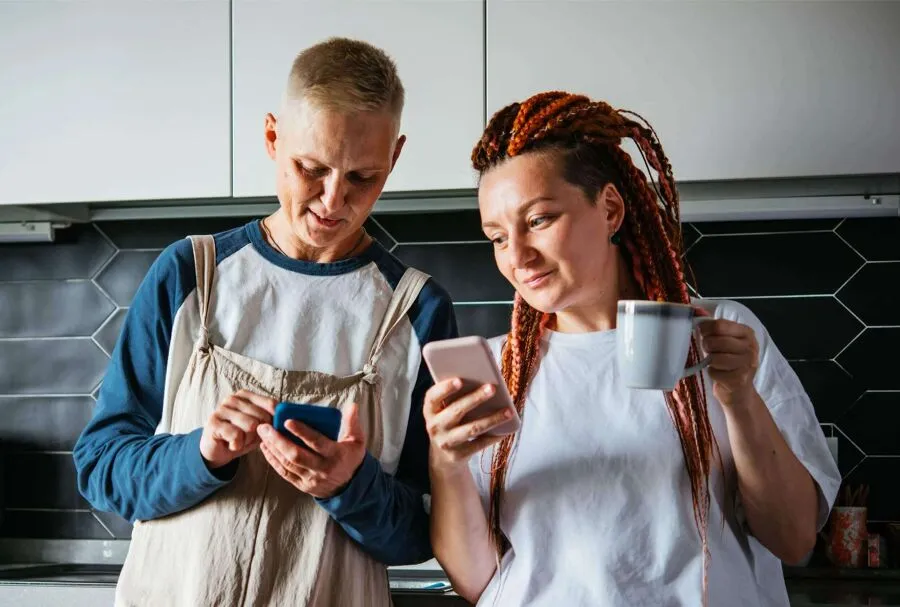 Two people are standing in the kitchen, looking at their phones.