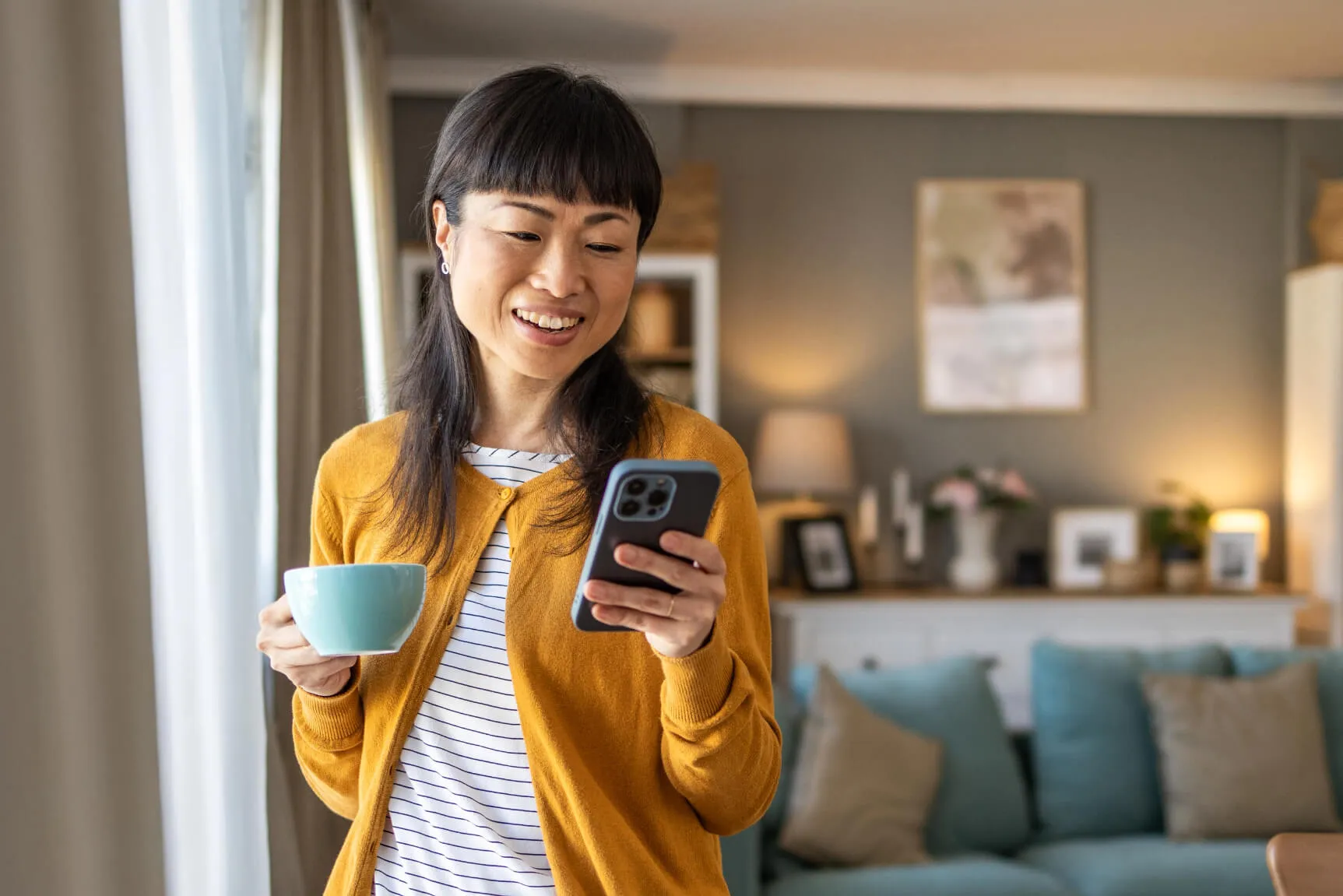 A woman smiling in a yellow cardigan holding a cup in one hand and her phone in the other