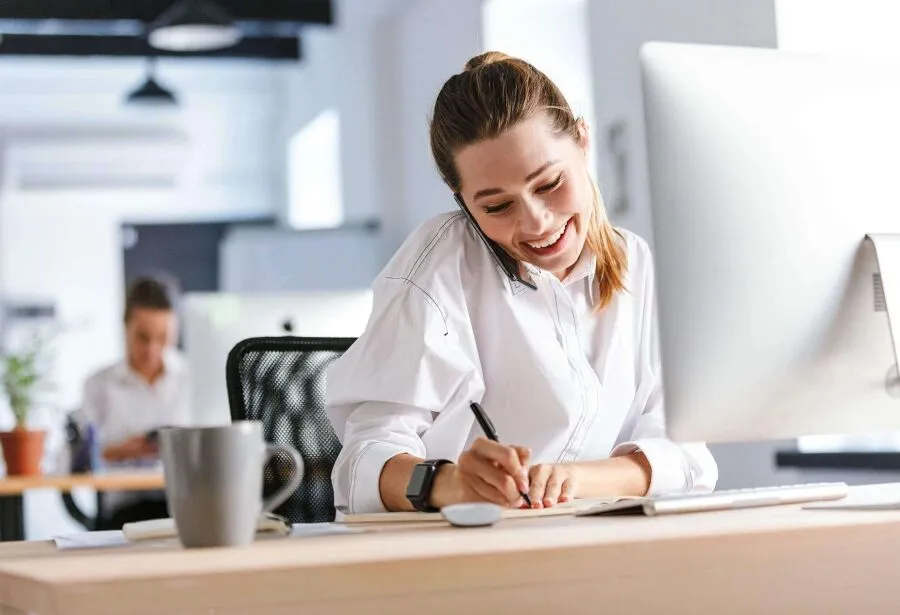 A woman sitting at desk in office, talking on phone and writing notes.