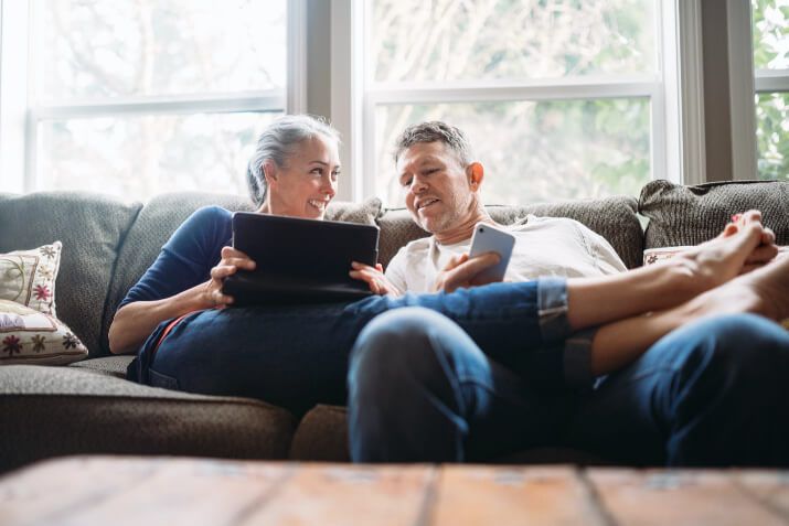 Smiling couple on a sofa. The woman shows the man something on her ipad whilst her legs lay across his knees and he is on his phone.
