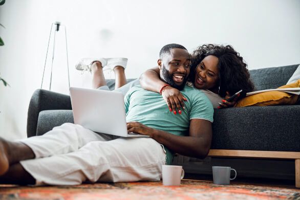 Woman laying on stomach on a sofa holding her phone with one arm around a man who is sat on the floor on his laptop, both are laughing