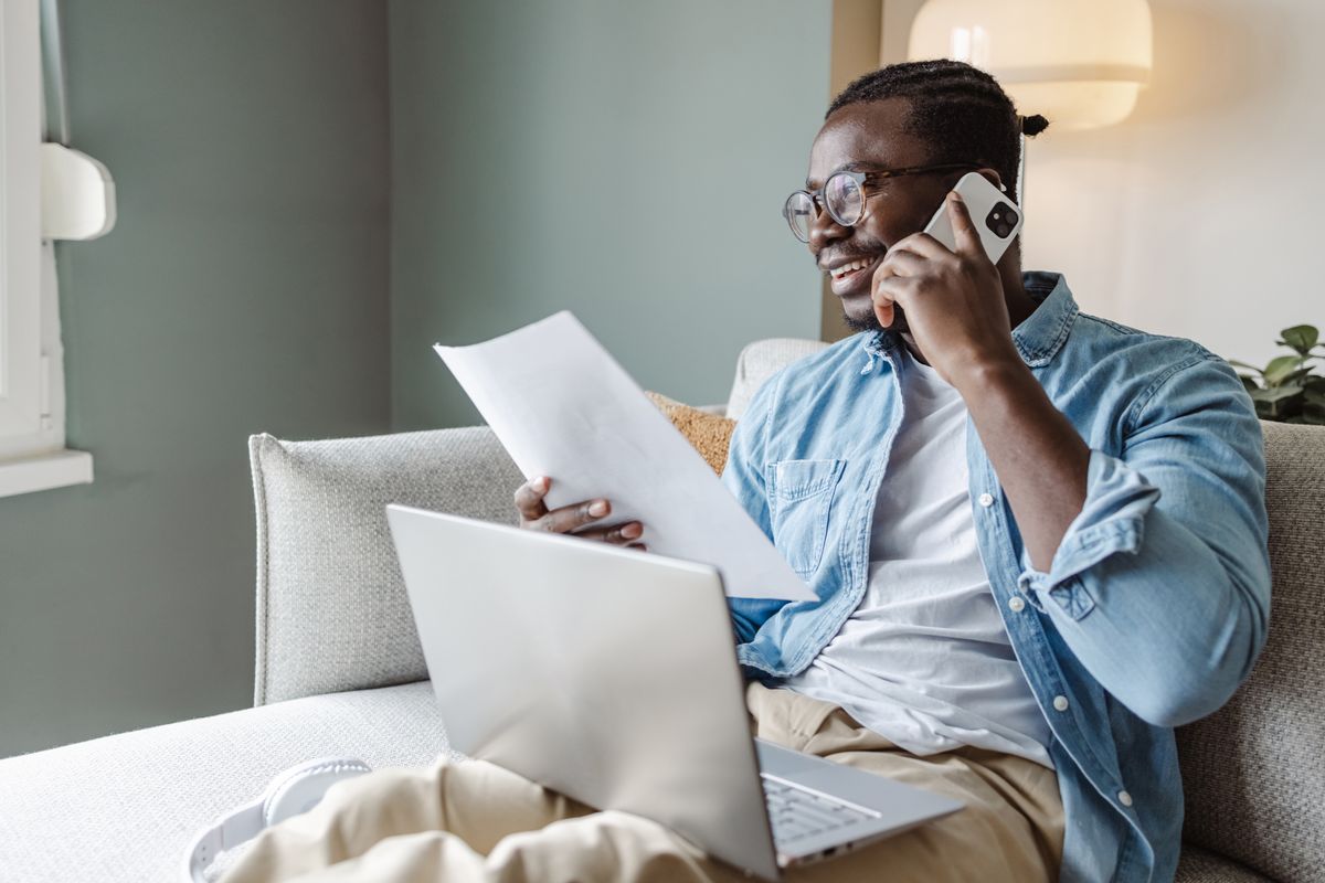 Bearded man with blue denim shirt is sat down on the phone whilst smiling and looking at a piece of paper